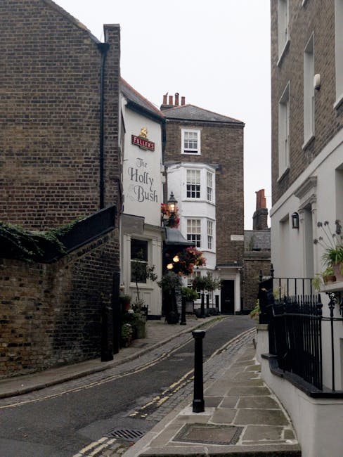 A narrow urban street scene showing a small, cobbled road flanked by tall, multi-story brick buildings with white window frames and decorative features. On the left, a building with a white sign reading 'The Holly Bush' is visible, with hanging flower baskets and potted plants along its exterior. The street has a slight incline and is bordered by short black metal bollards separating the roadway from the pedestrian pathway. To the right, a white building features a small balcony with potted plants and a black metal railing, and a wall-mounted light fixture is visible near the doorway. The scene is captured during daylight hours with natural light illuminating the buildings and street, highlighting the textures of the brickwork, cobblestones, and foliage, creating a quaint and charming atmosphere typical of historic West Hampstead areas in London relating to private on-site clearance or independent waste handling potential for local rubbish removal services.
