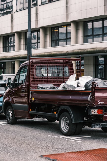 A small, dark red flatbed pickup truck parked on an urban street in front of a modern office building with large glass windows, loaded with various bags and loose rubbish items covered with black and white plastic sheets. The vehicle features a protective metal grille at the front of the cargo area, and its side panels and tailgate are clearly visible. The surrounding environment includes parked cars along the street, a street lamp pole positioned near the truck, and the building’s concrete and glass facade offering a contemporary backdrop. The scene captures the process of private waste collection, possibly linked to a rubbish removal service such as wasteclearancewesthampstead.co.uk, emphasizing the practical approach to on-site clearance for residential or commercial rubbish. The natural lighting highlights the textures of the rubbish bags and the truck's finish, providing a clear, professional view of the vehicle used for independent waste disposal in an urban setting.