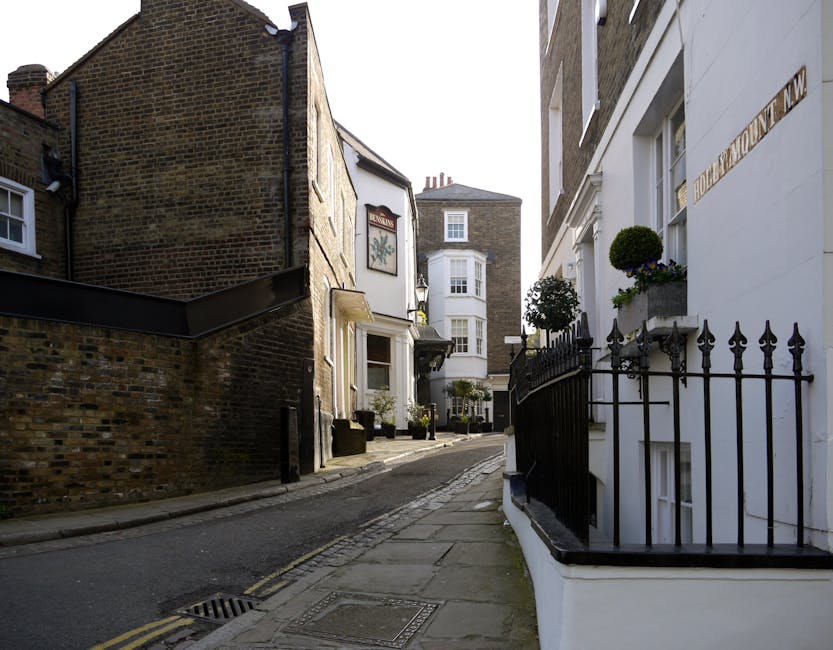 A narrow urban street scene with a gentle incline, showing a mix of residential buildings on either side. On the right, a white-painted building features a decorative black wrought iron fence with pointed finials, a small window with potted plants, and a white wall with gold lettering. To the left, a brick building with a textured, weathered appearance is partially visible, with a small bay window. Further ahead, a white building with a bay window and a sign displaying a clock or emblem is situated at the corner. The pavement is paved with concrete slabs, and the street has a drainage grate and a double yellow line along the curb. The environment suggests an inner-city or historic district area with a combination of old and modern architectural details, suitable for properties requiring private waste collection or on-site rubbish clearance with minimal disturbance to the street scene.
