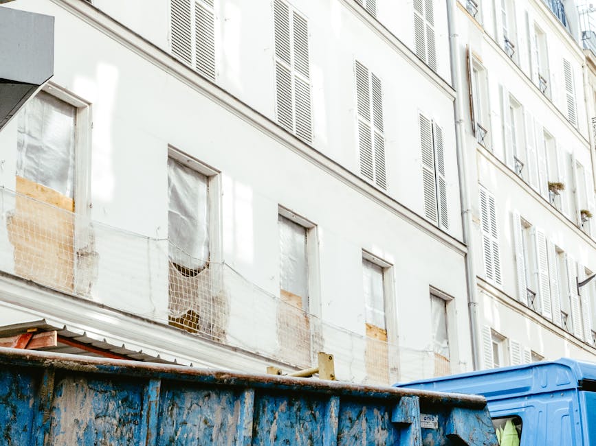 The image shows a section of a multi-storey residential building with white-painted facades and closed window shutters, some of which are partially covered with protective plastic sheeting or mesh. Below, a blue wheeled skip container is positioned on the street or paved area, filled with mixed waste materials including cardboard, wooden panels, and plastic debris, indicative of ongoing refurbishment or clearance work. The skip’s surface appears weathered with scratches and marks, contrasting with the clean building exterior. The scene is lit with natural daylight, with shadows cast from nearby structures, and the environment suggests an urban setting where private waste removal services, such as those provided by wasteclearancewesthampstead.co.uk, are utilized as an alternative to communal rubbish collection. The overall composition highlights the typical arrangement of waste management during building renovation or refurbishment in a residential context, with clear visual emphasis on the waste materials awaiting disposal or recycling.
