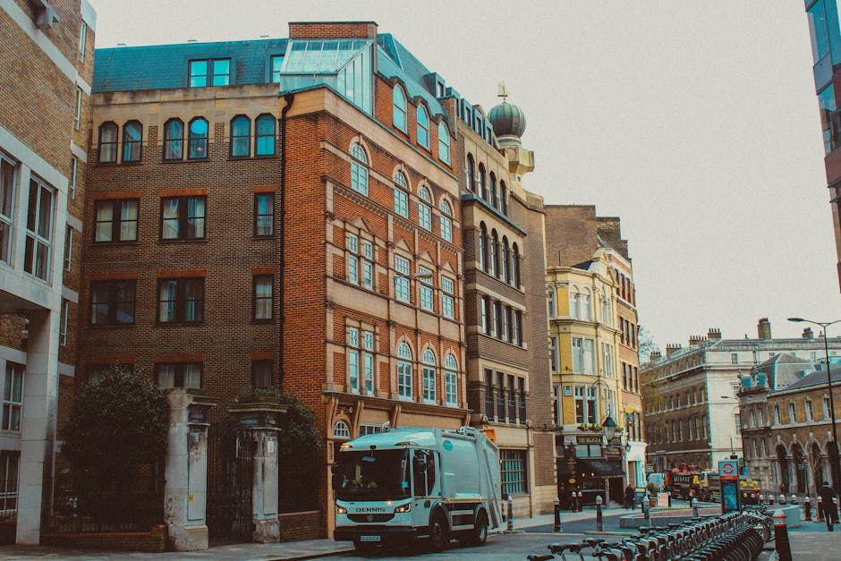 A street scene in an urban area featuring a mix of historic and modern buildings, with facades made of red brick, stone, and pastel-colored plaster. The foreground shows a light grey commercial waste collection vehicle parked on the pavement, partially blocking the view of the entrance to a building with wrought iron gates and stone pillars. The street is lined with metal bike racks, and a row of small bollards separates the sidewalk from the road. In the background, several other vehicles and a cityscape with multi-storey structures are visible under a slightly overcast sky. The scene captures the practical aspect of rubbish clearance in city environments, where private waste collection plays a key role alongside public disposal services, reflecting the context of alternative waste handling options for urban properties and businesses. The overall lighting is diffuse, contributing to a neutral, professional atmosphere typical of a day in the city focused on municipal and private waste management.