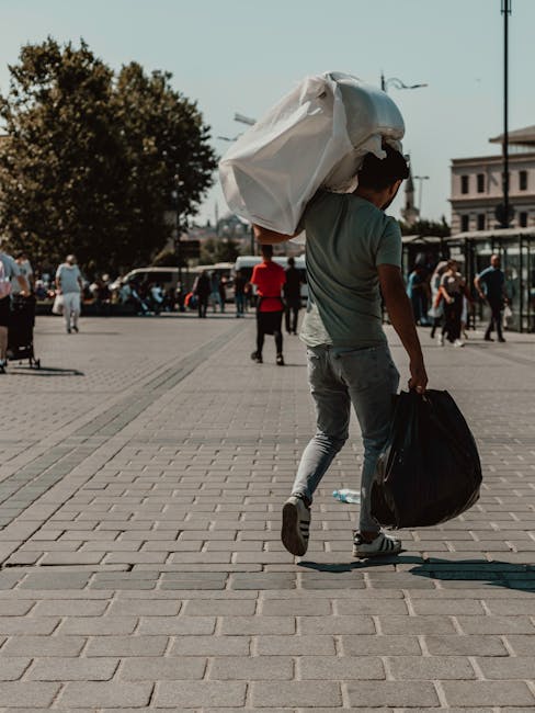 A young man walks across a paved urban square during daytime, carrying a large white plastic bag over his shoulder and holding a black garbage bag in his right hand. He is dressed in a light grey t-shirt and light blue jeans. The background features several pedestrians, some walking, some standing, and parked vehicles along the street, with a mix of trees and multi-story buildings visible. The scene is well-lit with natural daylight, and the pavement consists of rectangular bricks arranged in a grid pattern, typical for city walkways. This image, referencing independent waste handling and private rubbish collection, illustrates the act of on-site rubbish removal, supporting services like those offered by waste clearance companies such as wasteclearancewesthampstead.co.uk, in an everyday urban environment, emphasizing personal waste disposal distinct from municipal collection.
