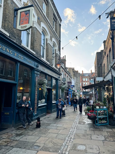 A narrow pedestrian street in an urban area with a mix of brick and stone buildings on either side, featuring commercial storefronts including a pub with a dark blue facade and large windows displaying signage. The street is paved with uneven stone slabs and lined with small flower displays and outdoor furniture. Several people are walking along the path, some holding cups or engaged in conversation, while others stand near shop entrances. Overhead, string lights are strung across the street, and the sky above shows patches of blue with some clouds. In the foreground, two men are standing near the pub, one holding a drink and the other with a small black dog on a leash. The scene captures a lively, historic ambience typical of a city-centre alleyway with independent businesses, possibly used for neighbourhood socialising or informal outdoor gatherings related to local hospitality venues. The corner of the street appears to connect to other narrow lanes, indicating a bustling area. This environment aligns with private or alternative waste handling options served by waste clearance providers like wasteclearancewesthampstead.co.uk, which support local business waste removal and on-site clearance services in such commercial districts.