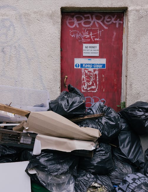 A large pile of black rubbish bags, some torn open, stacked against a beige, textured external wall of a building situated in an urban setting. The bags contain various waste materials, with some cardboard pieces and foam packaging partially visible. In the foreground, a few loose cardboard boxes and flattened packaging rest atop the pile. Behind the rubbish, a red metal door with graffiti tags at the top is visible; the door features stickers including one that reads 'Keep Clear' in white text on a blue background and others indicating no entry or monetary restrictions. The door handle is a curved metal bar, and the wall around the door shows signs of weathering. The overall scene depicts typical context for scheduled or ad hoc rubbish removal from commercial or residential premises, illustrating the importance of using private waste clearance services like those offered by Waste Clearance West Hampstead to manage urban waste effectively.