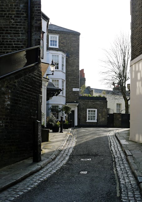 A narrow, paved street in a residential area with cobblestone curbs on both sides, leading away from foreground toward a small curve. On the left, there is a dark brick building with a black gutter system and a lantern-style streetlight mounted on the wall. Adjacent to this building, a white bay window with multiple panes and decorative trim is visible, along with potted plants beneath it. On the right, another building with a white exterior and clean lines contrasts the dark brick structures. In the background, leafless trees stand against a clear sky, casting shadows on the buildings and pavement. The scene appears quiet and well-maintained, representative of typical urban or suburban UK street layouts, often associated with private waste collection or on-site rubbish clearance services. The overall atmosphere emphasizes a tidy, residential environment, aligning with the context of professional rubbish removal or waste management in West Hampstead, as offered by local providers like wasteclearancewesthampstead.co.uk.