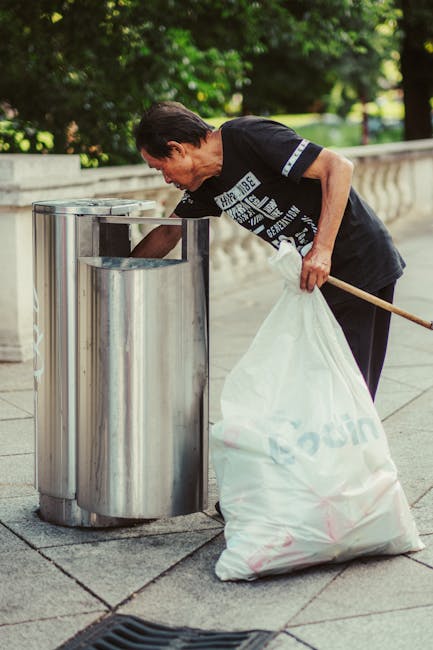 A middle-aged man with dark hair, wearing a black T-shirt with white text and dark trousers, is seen on a paved sidewalk, bending slightly to dispose of waste into a silver, cylindrical recycling bin with a rounded top. He holds a white plastic bag filled with rubbish in his left hand while using his right hand to insert waste into the open slot of the bin. The scene is outdoors, with lush green trees and foliage visible in the background, suggesting a park or urban green space setting. The pathway is made of light grey stone slabs, and a small black drainage grate is situated nearby. The image captures a moment of private waste disposal, consistent with onsite or independent rubbish collection practices, and relates to waste management in public areas as part of local or alternative rubbish handling services.